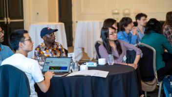 Diverse attendees attentively listening at round conference tables with laptops and projector.