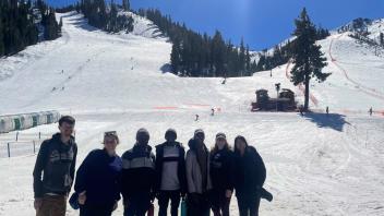 Group of people standing on snowy ski slope beneath sunlit blue sky and tree-lined ridge