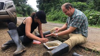 Two people in rubber boots sit on the side of a dirt road pipetting muddy water from a shallow tray.