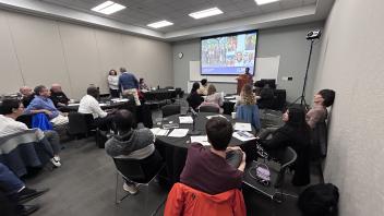 A conference room of people at tables watch a presentation on a large screen