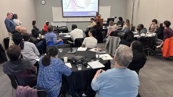 A conference room of people at tables watch a presentation on a large screen