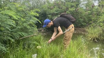 Two people crouching at a grassy marsh edge, reaching into a small pond under cloudy sky