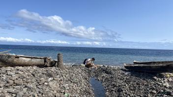 Two people on rocky shore by driftwood, stream to blue ocean beneath clear sky