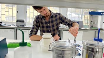 Young man in plaid shirt adjusting glassware beside metal canisters and plastic bottles in lab