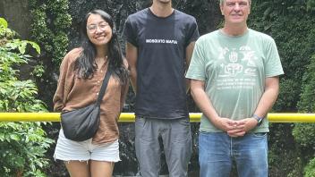 Three smiling people standing on a path in front of a small waterfall and greenery