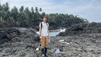 Man standing on black volcanic rocks with backpack and gear, palm trees and ocean behind