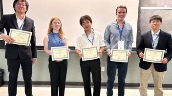 Four people stand in the front of a lecture hall holding paper certificates and smiling.