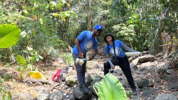 Two hikers in blue shirts pose on rocks in a shaded jungle stream with large green leaves