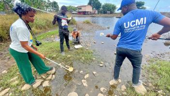 Volunteers cleaning trash from a shallow, rocky stream near homes on a sunny day