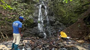 Narrow ravine waterfall with two people (blue and yellow jackets) on black pebble shore, photo