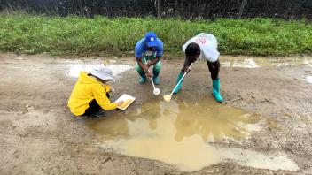 Three people in rain gear kneel on a muddy road, scooping water from a puddle.