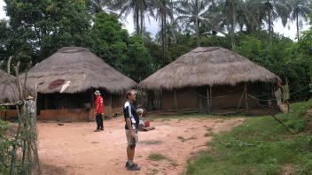 A few people stand around an open dirt area in front of two straw roof houses