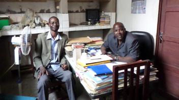 Two people in a small office sit on opposite sides of a desk filled with papers.