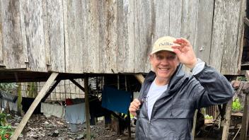 A man waves to the camera standing next to a house built on stilts with laundry drying underneath