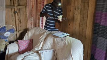 A man with a mouth aspirator and a net-covered cup looks for mosquitoes behind two chairs in a home