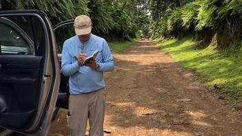 A man stands next to the open doors of a car along a dirt road. He is writing in a small journal