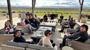Many people seated in lounge chairs sit outdoors under a shade in front of a vineyard