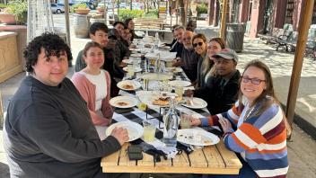 Many people sit at a long table outdoors with pizzas on the table.