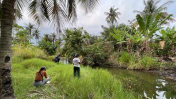 Two people by a grassy tropical stream with palm trees and an overcast sky