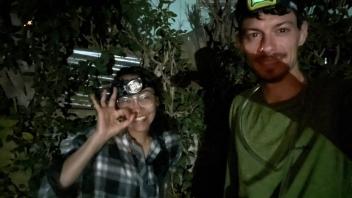 Two smiling people wearing headlamps in dark foliage, one making OK sign (photograph)