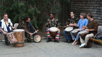A group of people play traditional drums in traditional dress sitting outdoors.