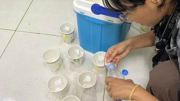 Person kneeling on tile pouring water from bottle into several white cups by a blue cooler.