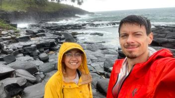 Two smiling people in yellow and red rain jackets on basalt shore with waves, palm trees (photo)