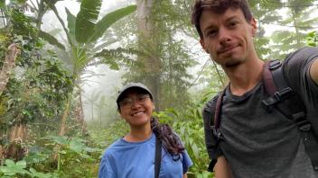 Two smiling hikers taking a selfie in a misty tropical forest with large green leaves