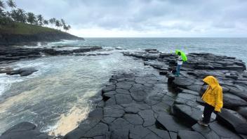 Two small people in yellow and green rain jackets on black basalt shoreline under gray sky