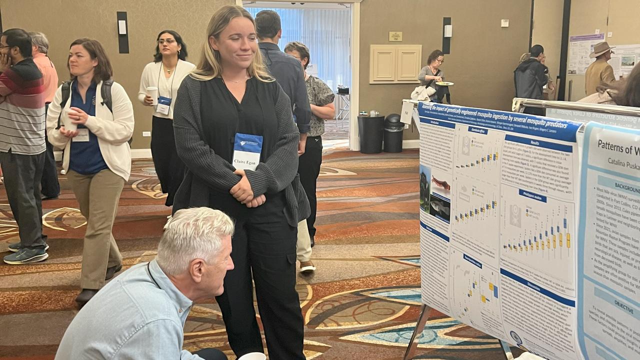Claire stands in front of her poster while an older gentleman crouches to read it better