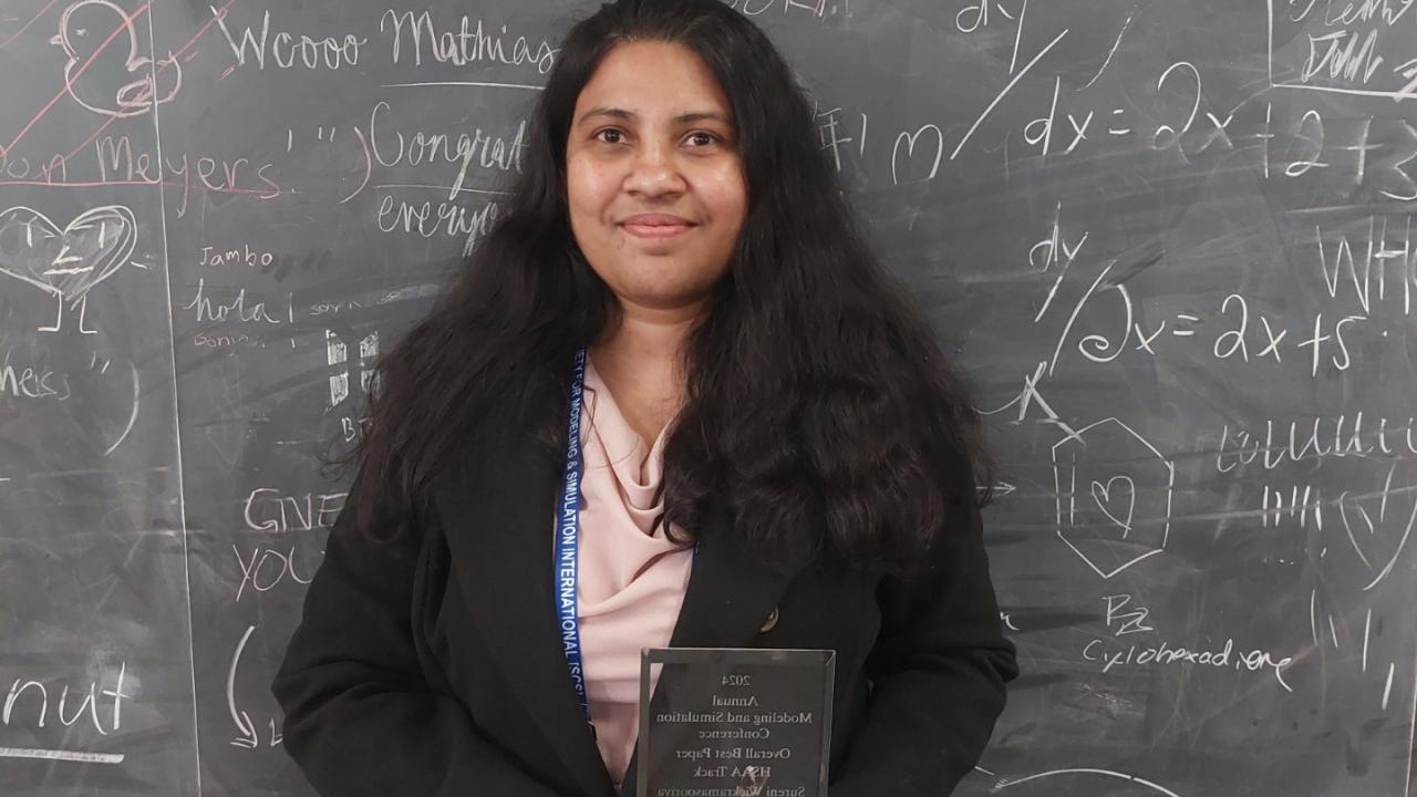 Sureni stands in front of a blackboard covered in mathematics and notes holding an etched glass award