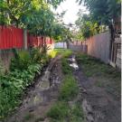 Muddy rutted dirt lane with puddles between red metal and wooden fences, leafy trees