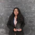 Sureni stands in front of a blackboard covered in mathematics and notes holding an etched glass award